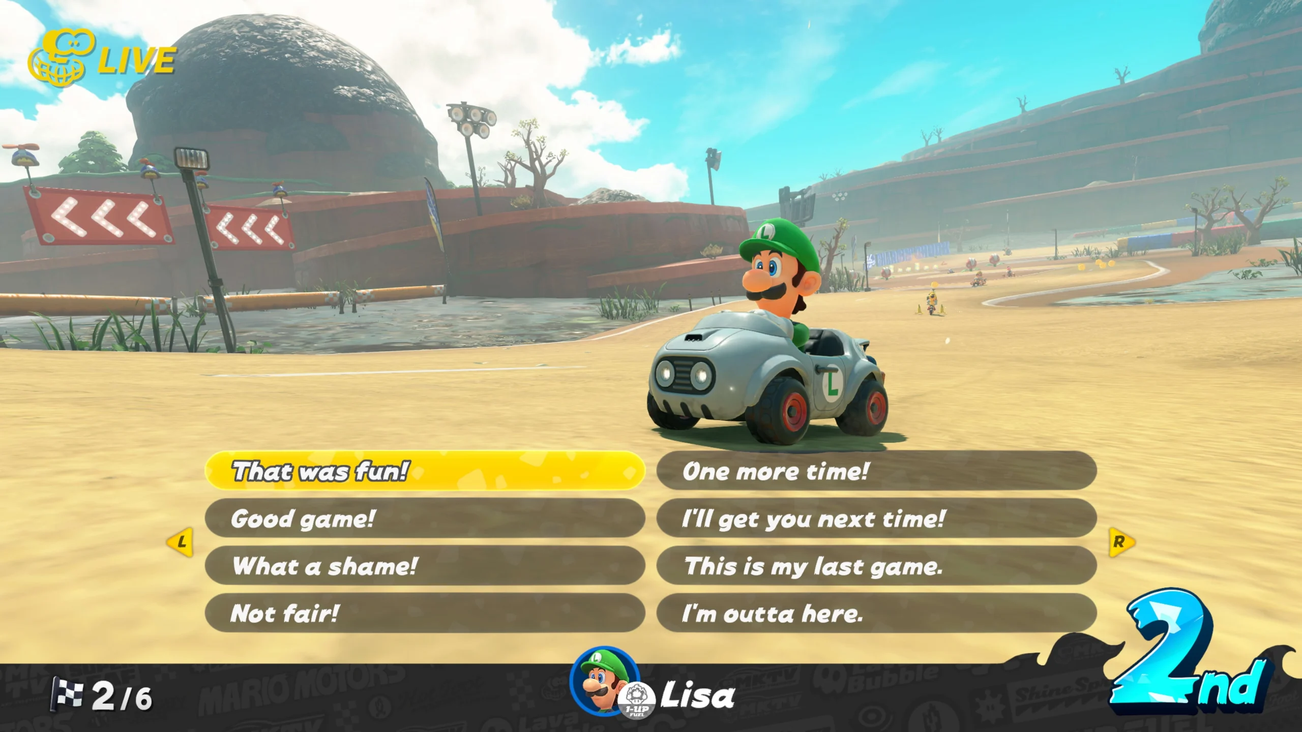 Luigi driving a kart on a desert track in Mario Kart World, displaying in-game chat options and 2nd place ranking