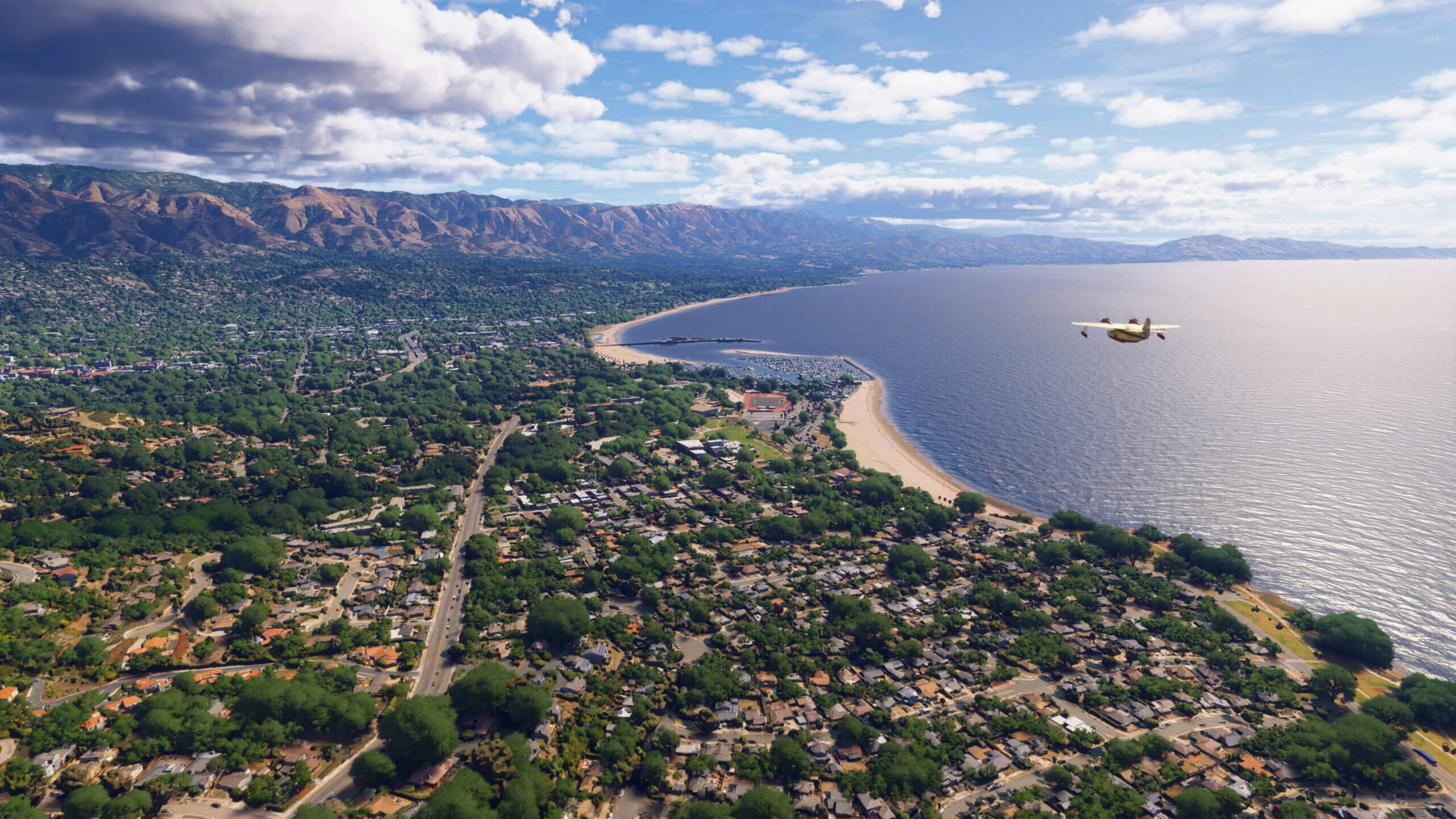 Aerial view of a coastal California city with lush greenery, mountains, and a plane flying over the ocean