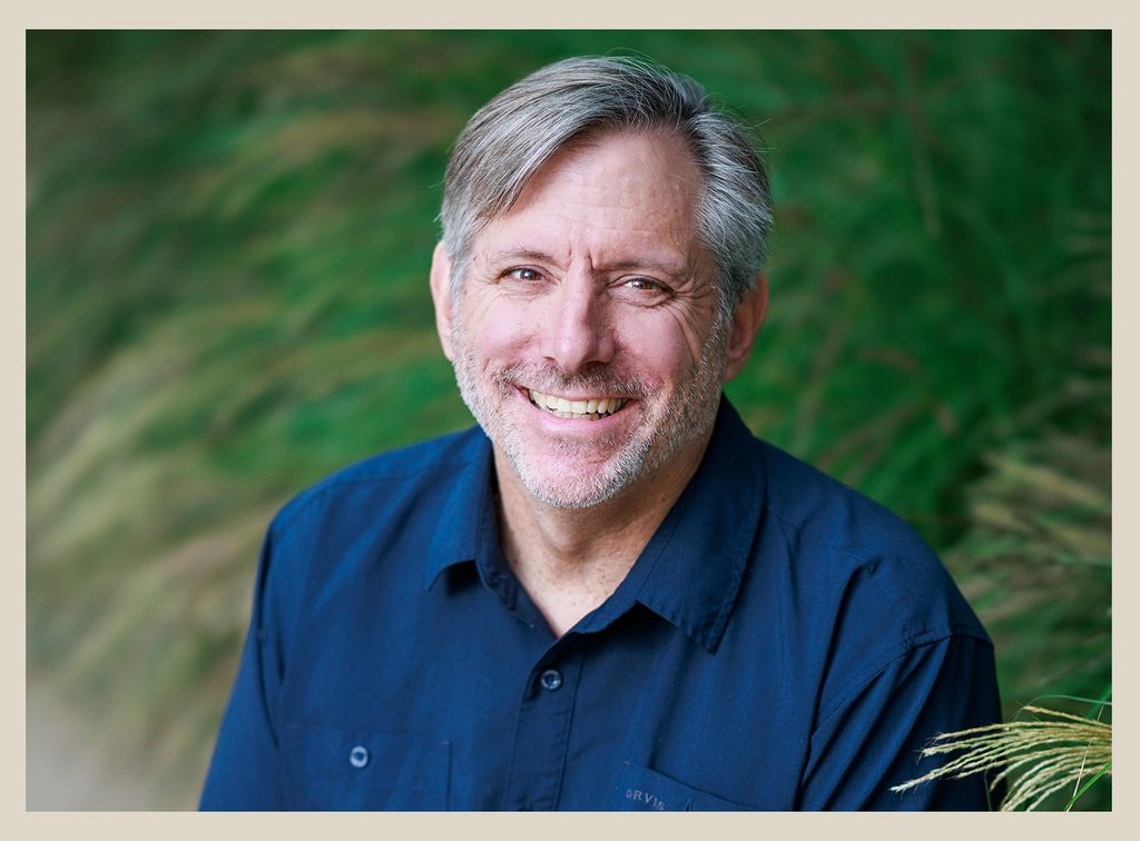 Smiling man with gray hair and beard in a blue shirt, standing in front of lush green foliage