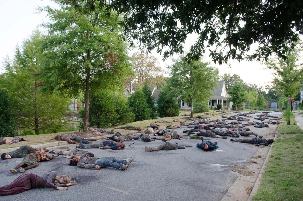 Street littered with fallen Walkers in a suburban neighborhood, trees and houses in the background