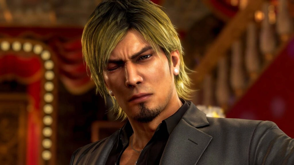 A serious-looking man with blond hair and a goatee, wearing a suit, stands in a dimly lit room