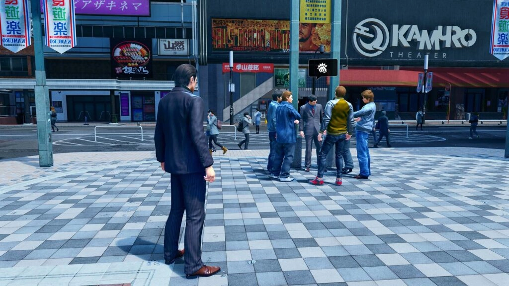 A man in a suit observes a group of people gathered outside Kamuro Theater in a bustling city setting