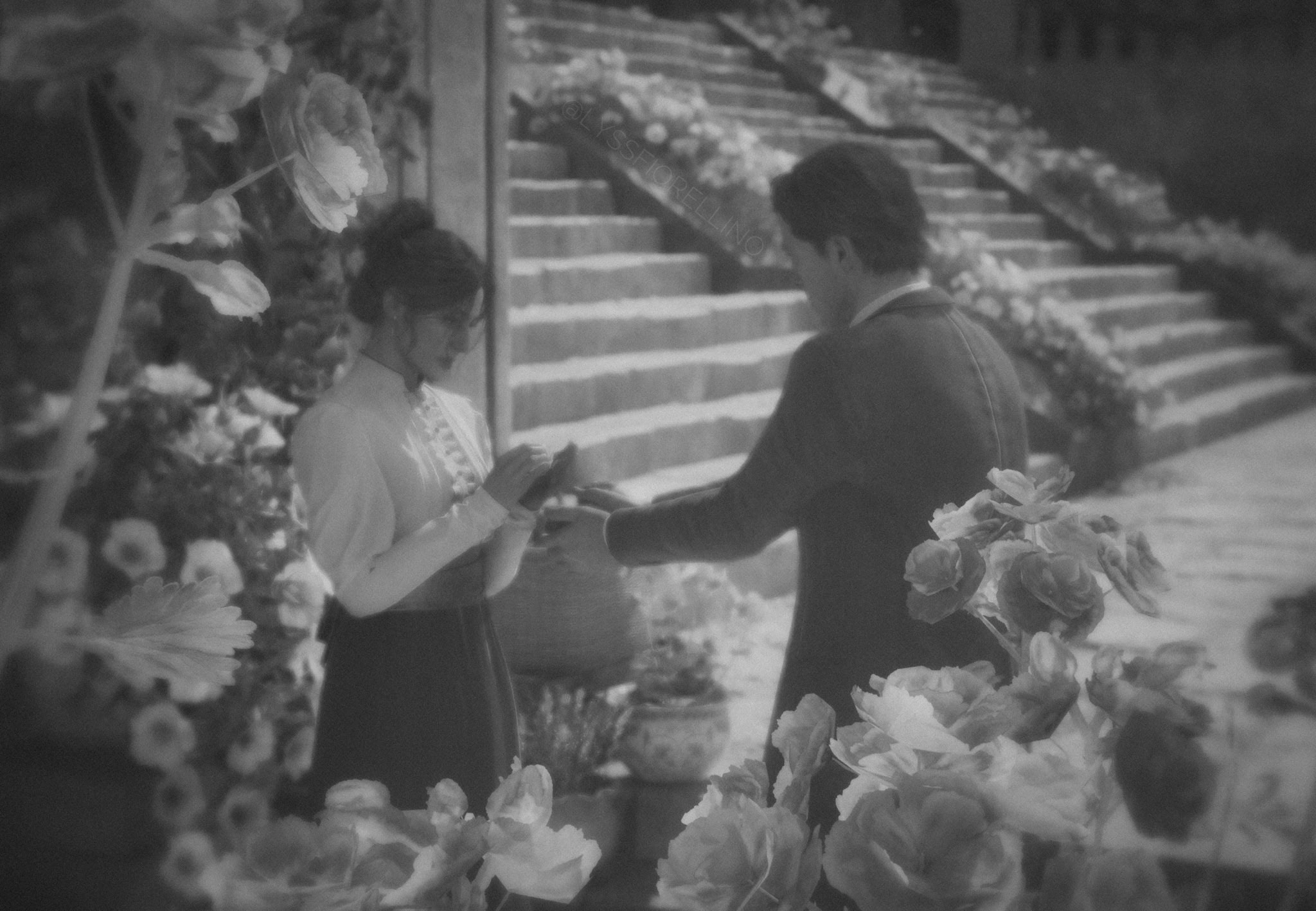 A couple exchanges rings in a romantic garden setting, surrounded by blooming flowers and stone steps