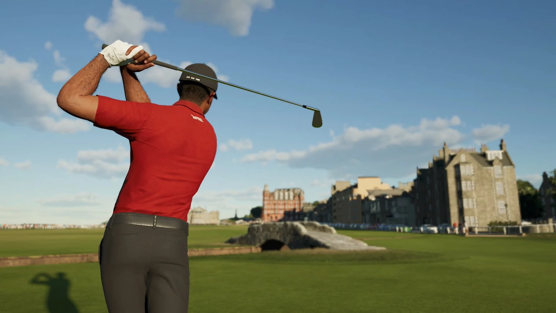 Golfer in red shirt swings club on lush course, historic buildings in background under clear sky