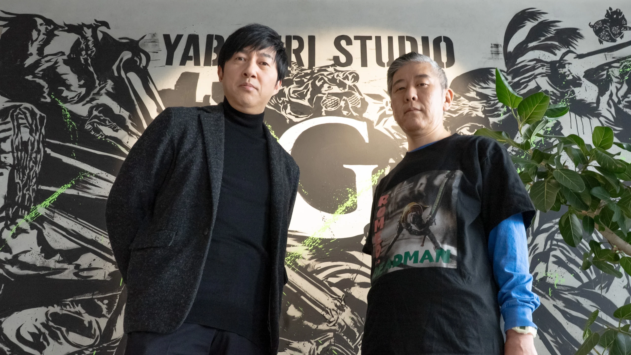 Two men standing in front of a Yabuki Studio sign, one wearing a black jacket, the other a graphic t-shirt
