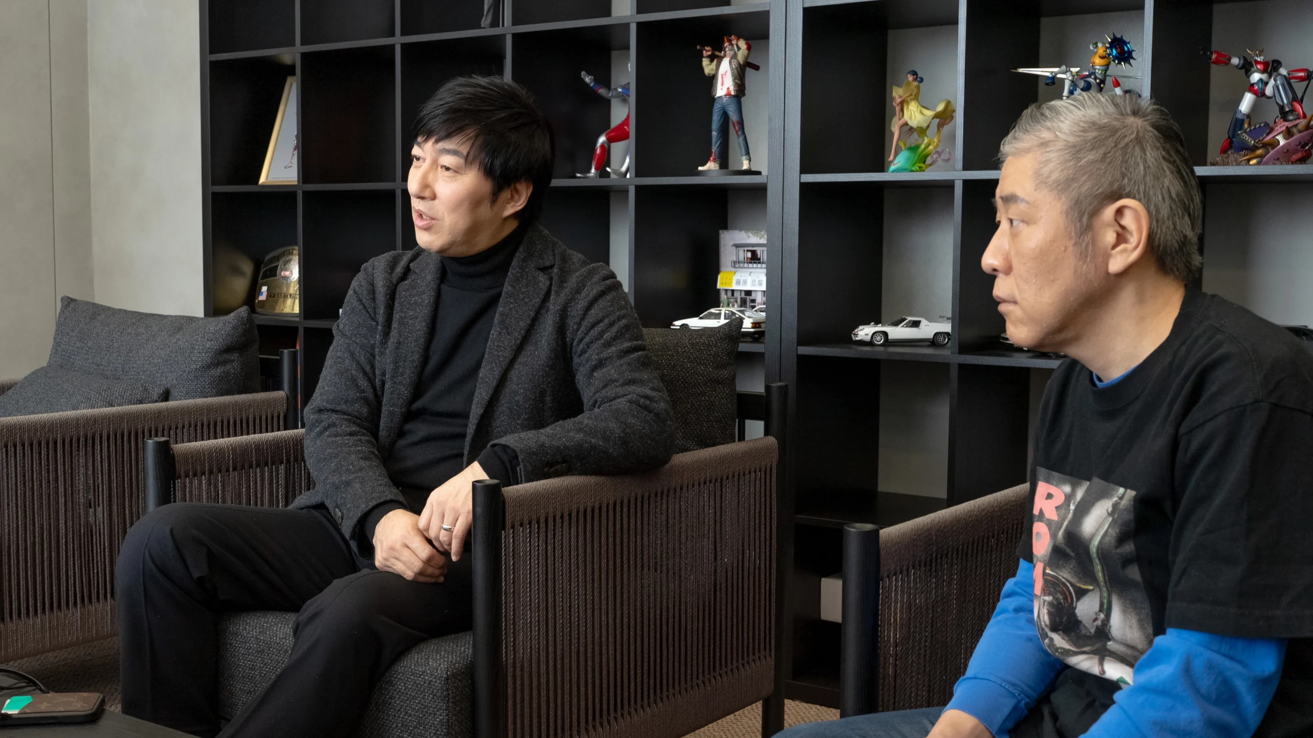 Two men sitting in chairs, discussing in front of a shelf with various action figures and collectibles