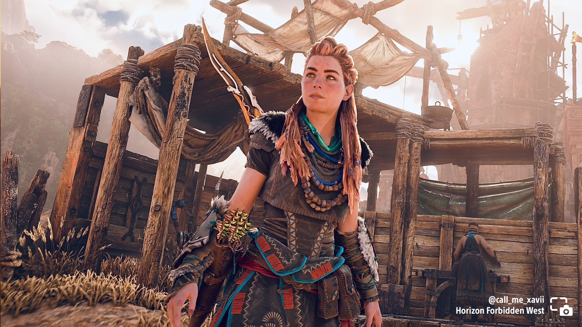 Aloy stands outside a rustic encampment in Horizon Forbidden West, showcasing vibrant tribal attire